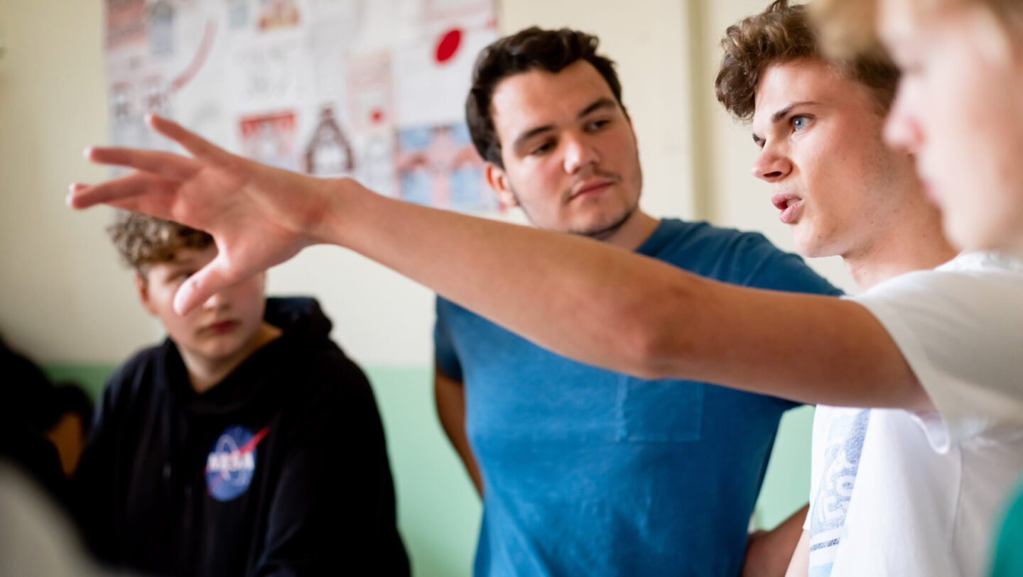 Schüler stehen in einer Gruppe und unterhalten sich. Ein Junge hebt seine Hand und erklärt etwas.