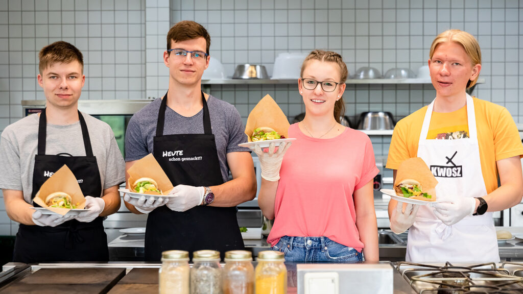 Vier Jugendliche stehen in einer Küche. Sie halten die Produkte ihrer Schülerfirma in der Hand - selbstgemachte Burger.