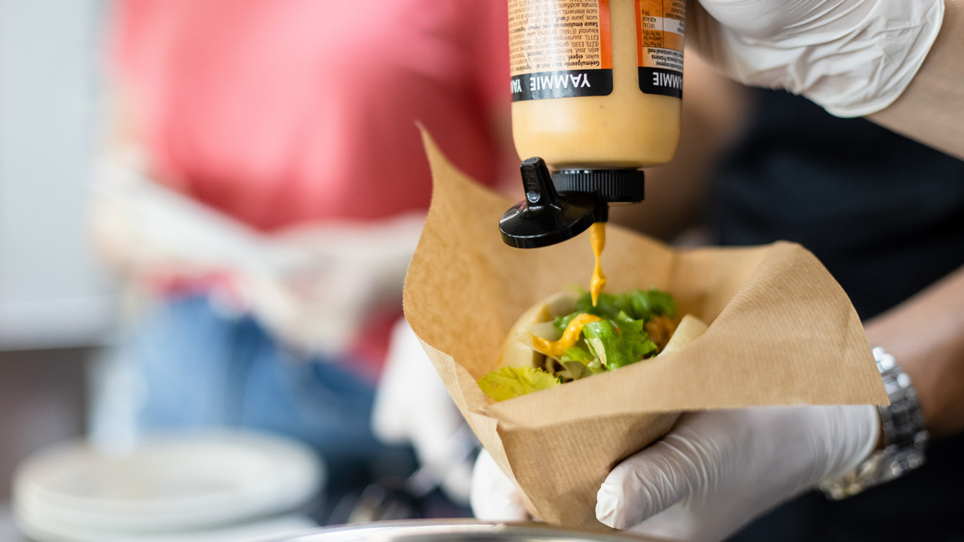 Eine Hand hält einen Burger auf den Sauce aus einer Tube getropft wird.