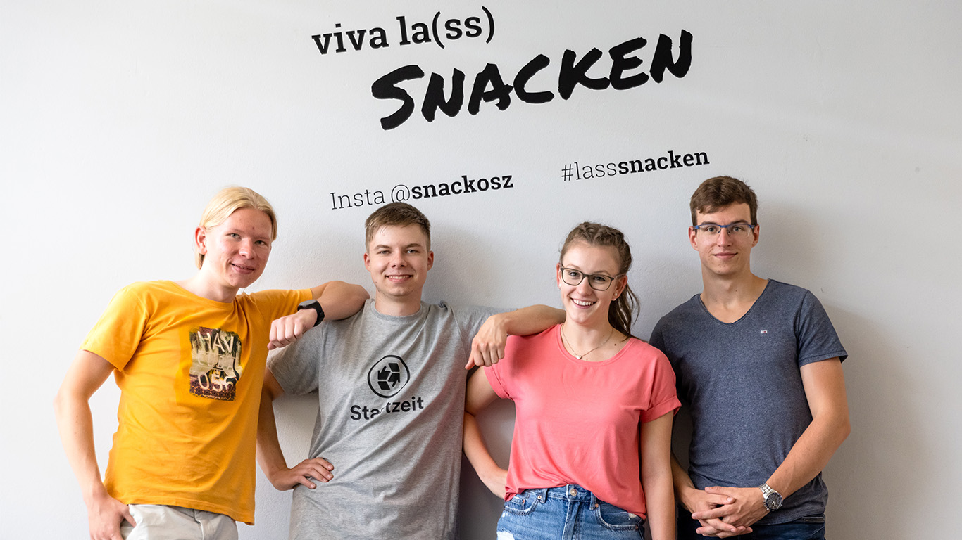 Vier Jugendliche stehen vor einer Wand mit der Aufschrift "viva la(ss) Snacken" Sie haben zum Teil ihre Ellenbogen auf die Schultern der anderen gelegt.