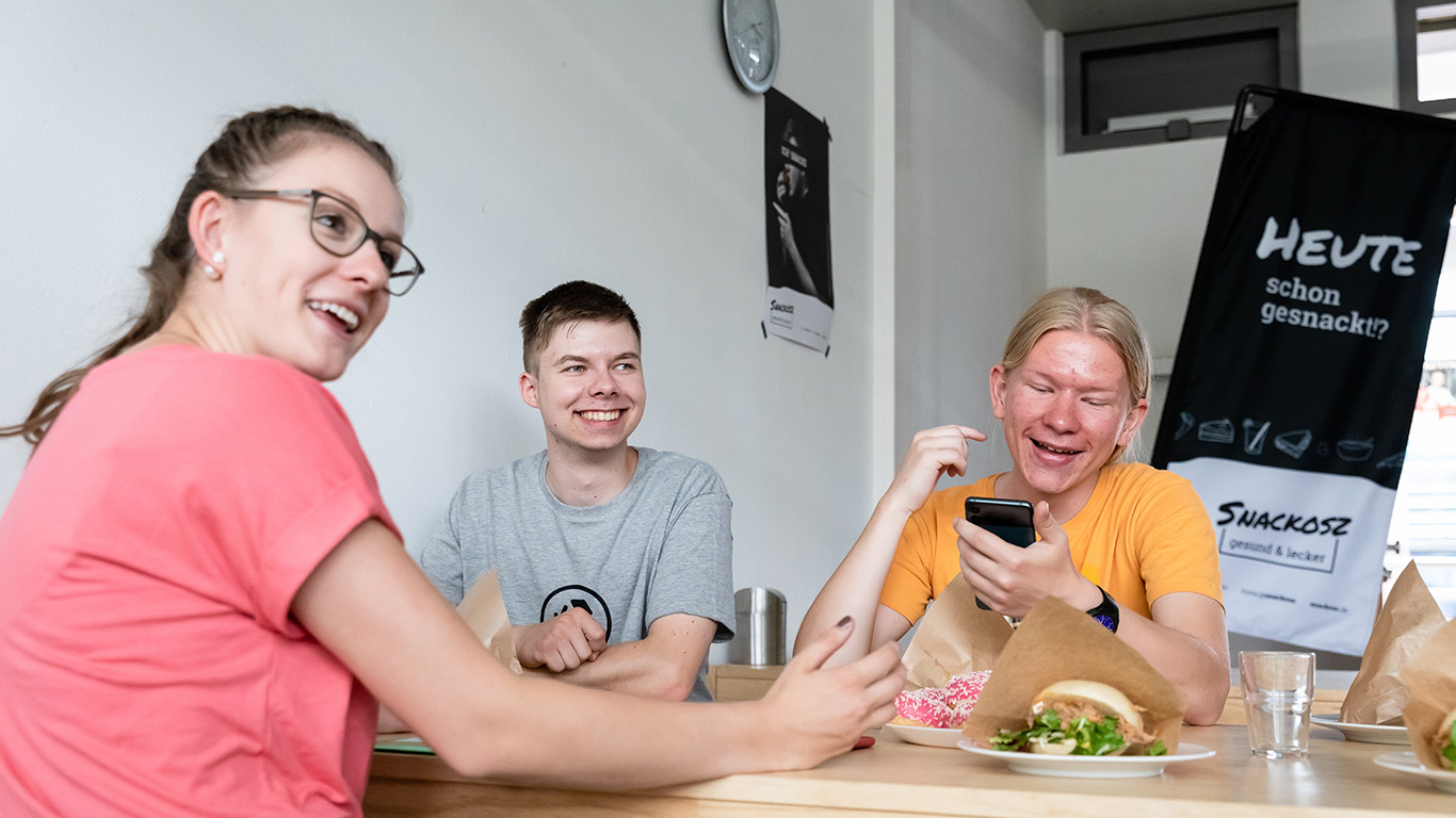 Drei Jugendliche sitzen an einem Tisch auf dem Essen steht. Sie lächeln.