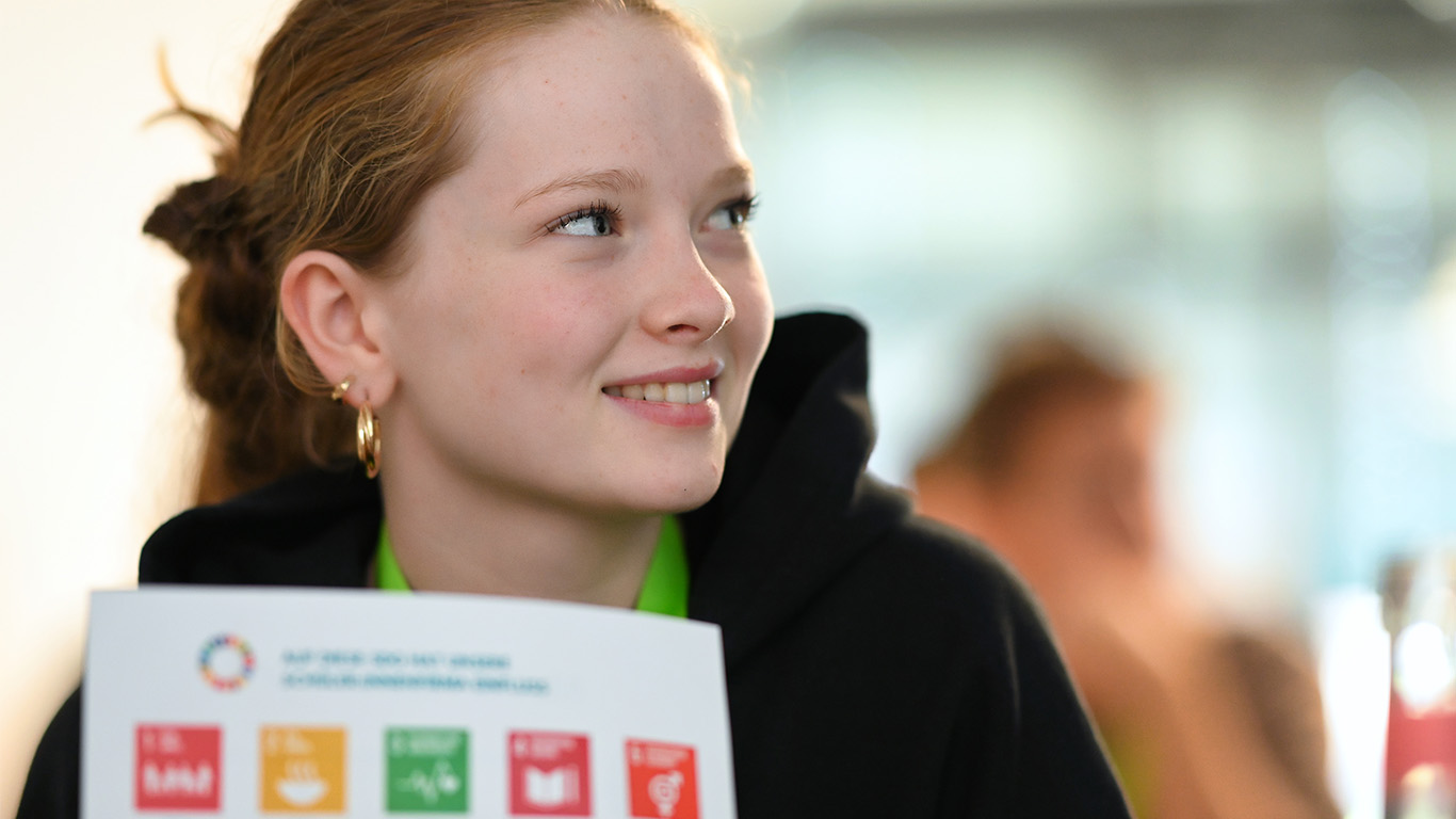 Ein Mädchen schaut zur Seite. Sie hat ein Papier in der Hand auf dem die Sustainable Development Goals zu sehen sind.