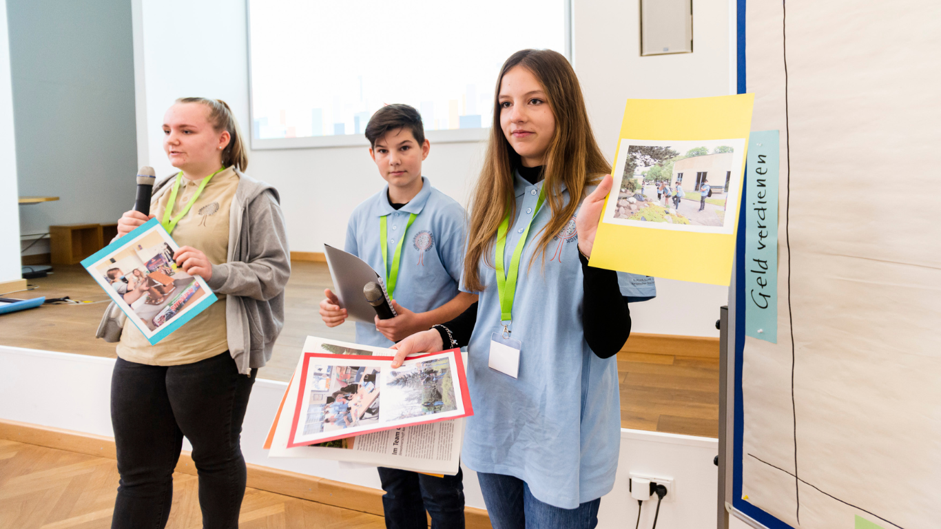 Drei Jugendliche präsentieren vor dem Publikum ihre Schülerfirma. Sie haben bunte Blätter dabei, auf denen sie Fotos aufgeklebt haben.