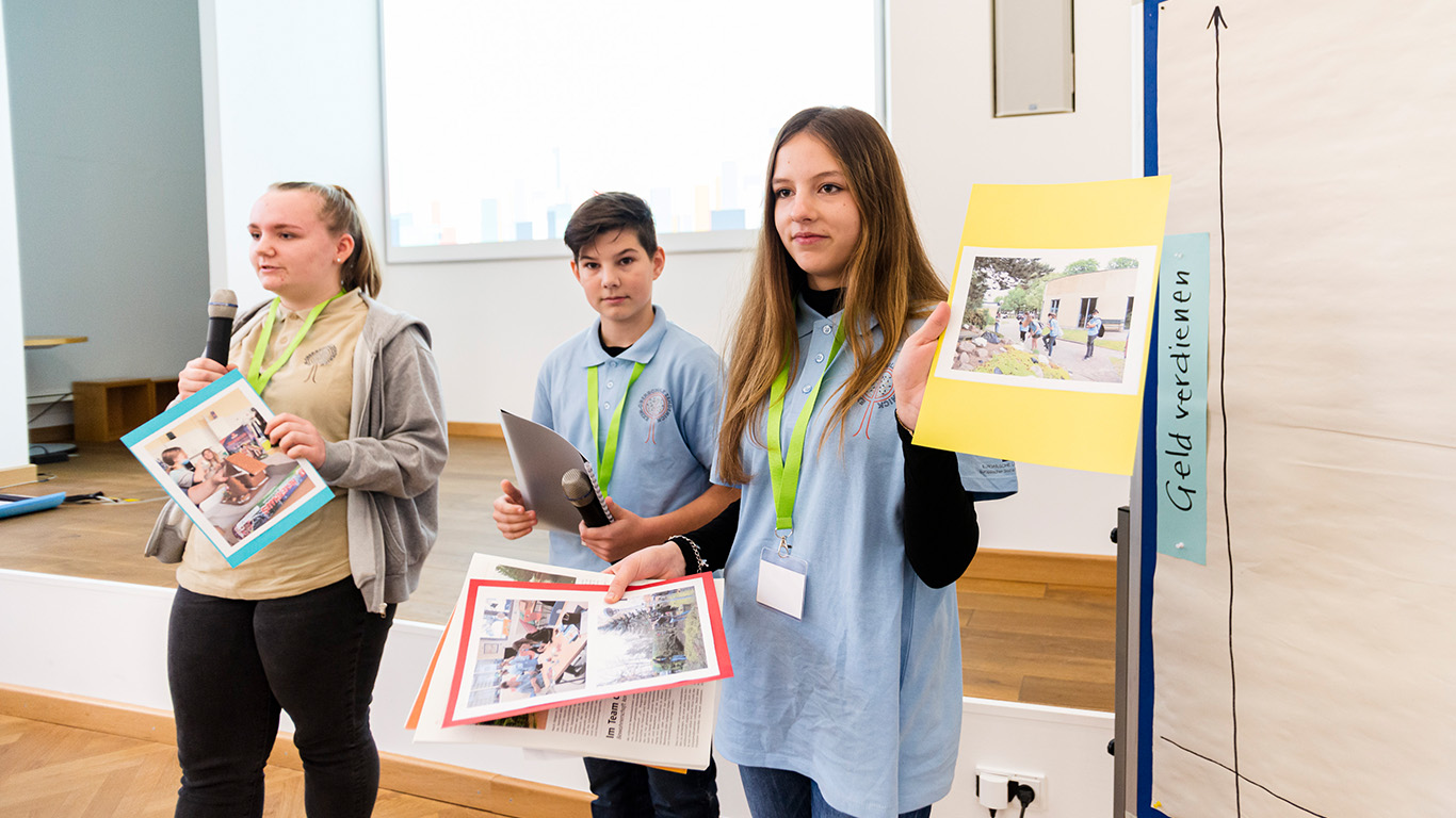 Drei Jugendliche präsentieren vor dem Publikum ihre Schülerfirma. Sie haben bunte Blätter dabei, auf denen sie Fotos aufgeklebt haben.