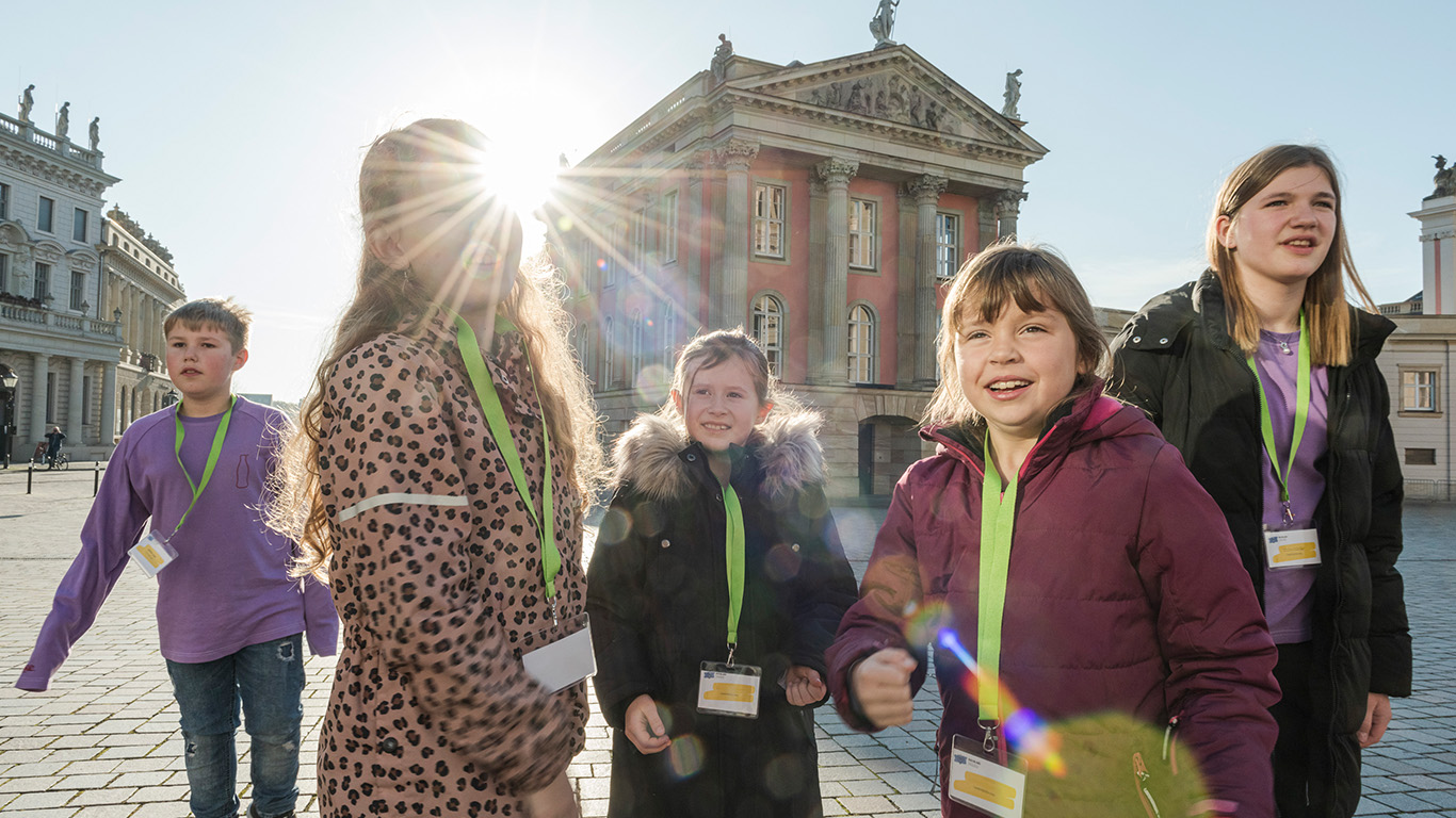 Kinder und Jugendliche stehen draußen auf einem Platz und lachen. Die Sonne scheint in die Kamera.