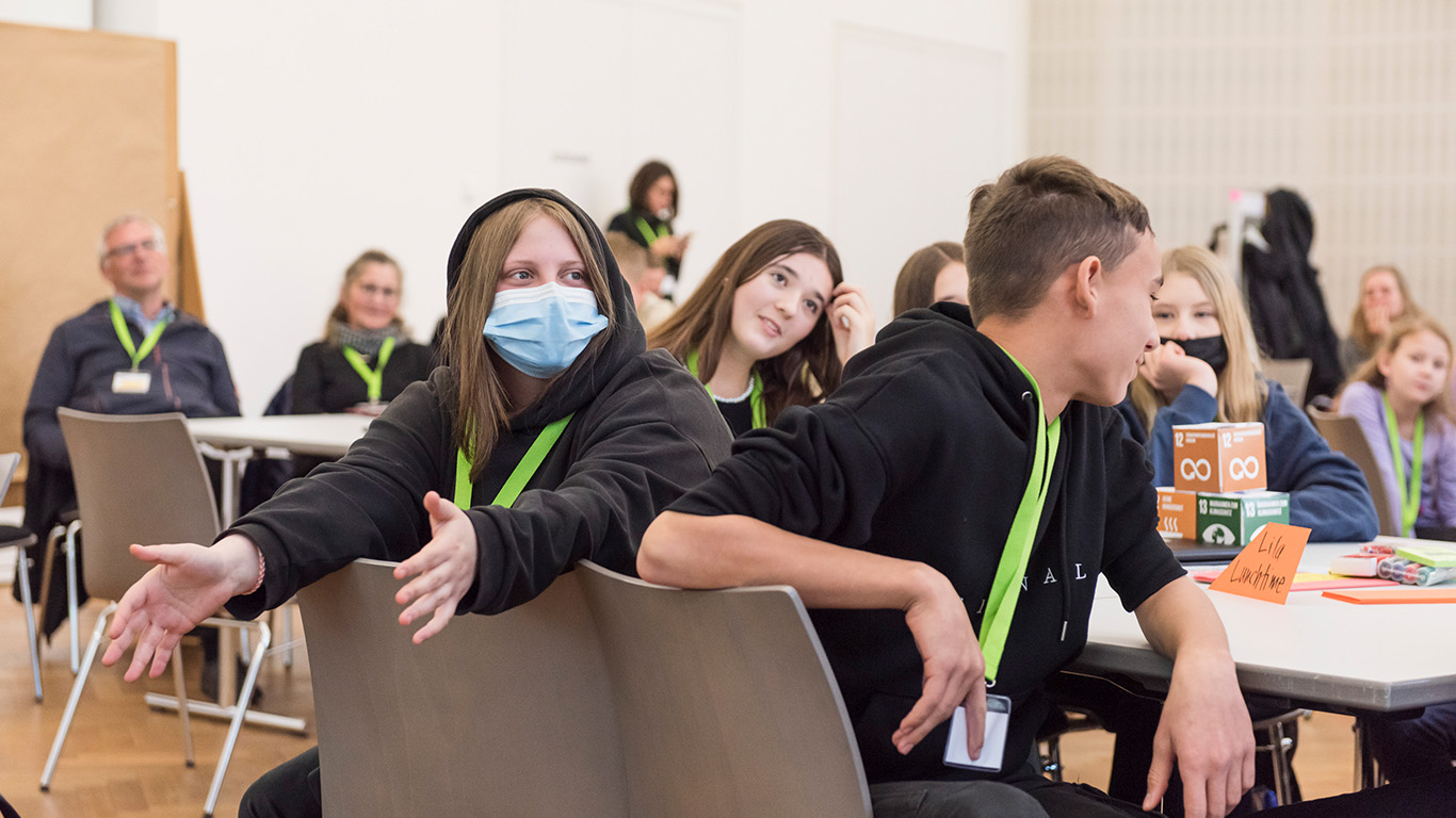 Jugendliche sitzen an einem Tisch und unterhalten sich. Sie tragen grüne Schlüsselbänder um den Hals.