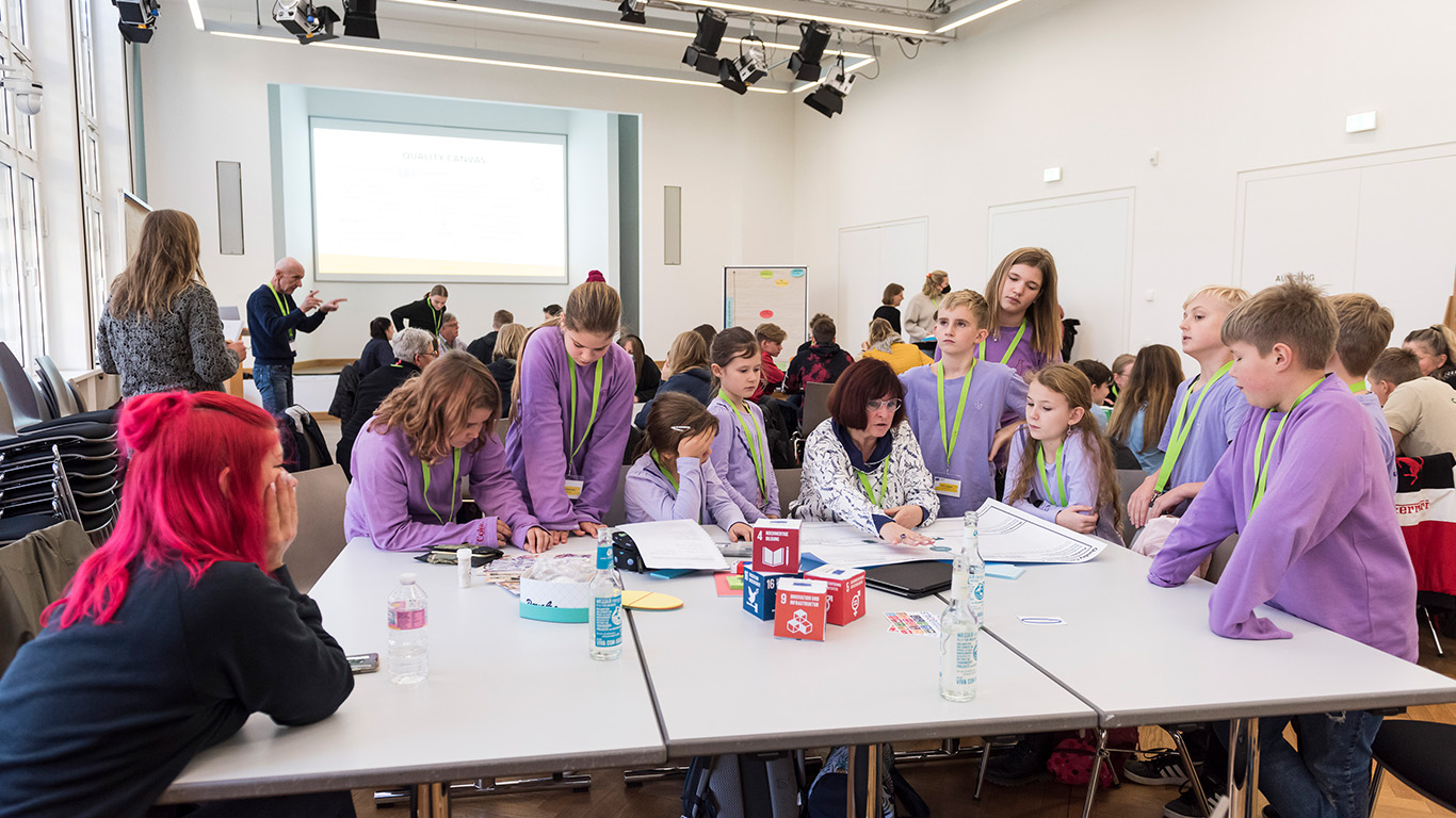 An einem Gruppentisch sitzen Kinder in lilafarbenen Pullovern und unterhalten sich über die Aufgabe.